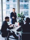 Geschäftsmann im formellen schwarzen Anzug, der neben seinen Kollegen steht und einen Finanzbericht während einer Konferenz in einem modernen Arbeitsraum analysiert und über die Implementierung der SAP Sales Cloud redet